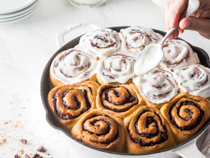 Mexican Chocolate Cinnamon Rolls in Cast Iron Pan with Spoon Pouring Frosting Over Top
