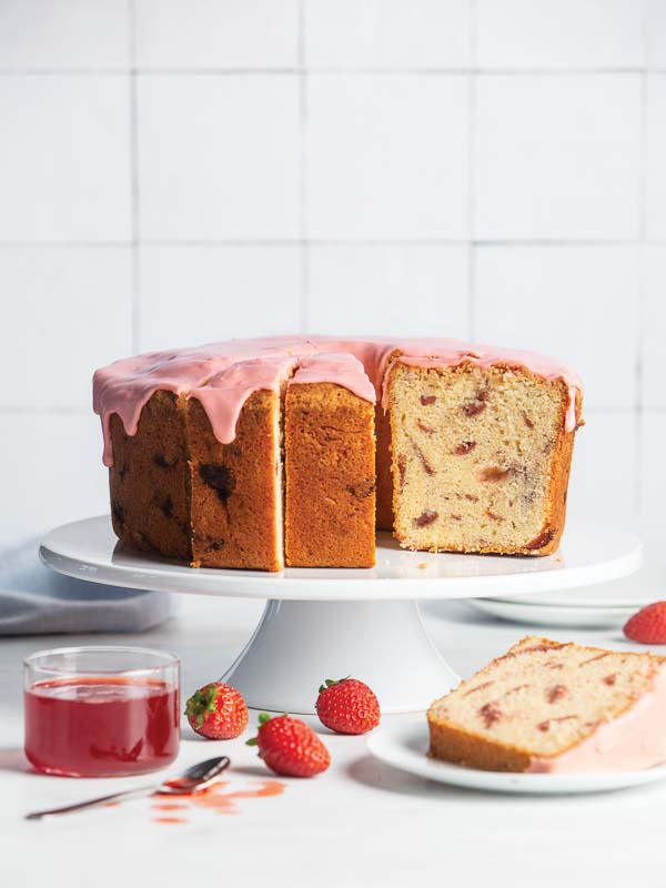 Roasted Strawberry Pound Cake on white cake stand with one slice of cake on white plate with strawberry jam with spoon and strawberries